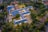 An aerial view of a large, L-shaped building complex surrounded by trees and pathways. The rooftops are blue, and the building is illuminated by outdoor lighting. The surrounding area includes other smaller buildings, open spaces, and residential houses.