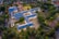 An aerial view of a large, L-shaped building complex surrounded by trees and pathways. The rooftops are blue, and the building is illuminated by outdoor lighting. The surrounding area includes other smaller buildings, open spaces, and residential houses.