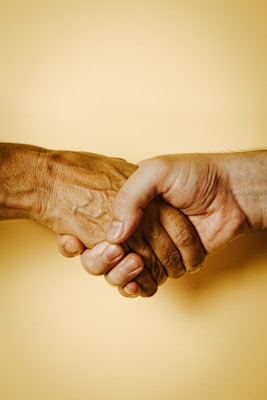 Two hands are clasped together in a handshake against a plain background. One hand appears older with visible wrinkles, while the other looks younger and smoother.