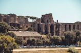 Ancient ruins with large arched structures stand surrounded by trees and a residential building in the foreground. The scene is set under a clear blue sky, creating a contrast between the historical architecture and the natural elements.