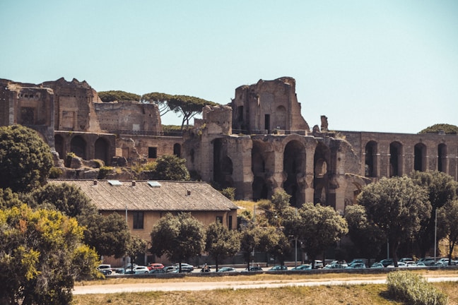 Ancient ruins with large arched structures stand surrounded by trees and a residential building in the foreground. The scene is set under a clear blue sky, creating a contrast between the historical architecture and the natural elements.