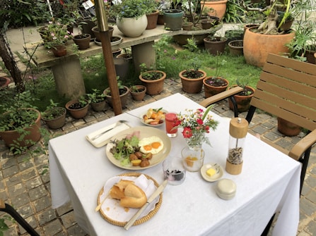 A table set for a meal in a garden setting, featuring a variety of dishes including a plate with fried eggs, potatoes, and salad, a baguette on a small plate, and various condiments. A vibrant red drink and a small vase with flowers add color to the scene. The background contains numerous potted plants and lush greenery, contributing to a relaxed and natural atmosphere.