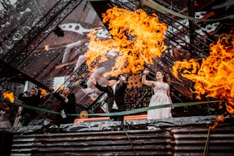 Several performers on stage are surrounded by dramatic flames, with a backdrop of intricate, geometric lighting. The performers appear to be singing passionately, and the intensity of the fire creates a dynamic and energetic atmosphere.