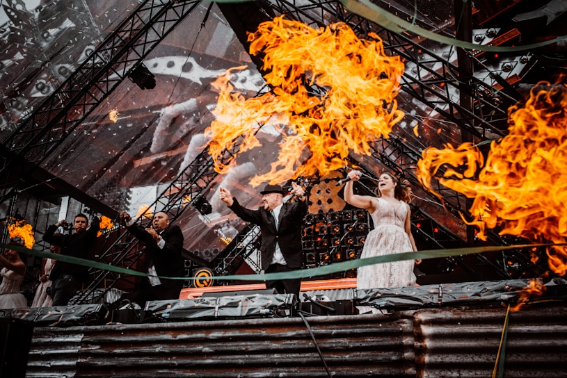 Several performers on stage are surrounded by dramatic flames, with a backdrop of intricate, geometric lighting. The performers appear to be singing passionately, and the intensity of the fire creates a dynamic and energetic atmosphere.