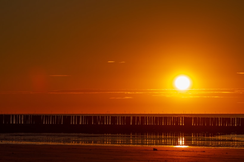 A vibrant sunset over a calm beach with the sun low on the horizon, casting a warm orange glow across the sky and water. The silhouette of wooden posts or pylons stretches across the length of the shoreline, reflecting softly in the wet sand.