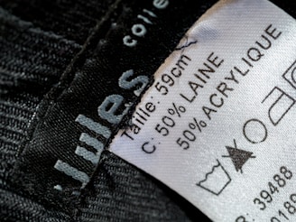 A close-up view of a clothing label showing washing instructions and material composition. The label states the size as 59 cm and the material as 50% wool (laine) and 50% acrylic. Laundry symbols are visible, including one indicating a prohibition against bleaching.