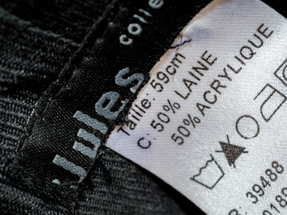 A close-up view of a clothing label showing washing instructions and material composition. The label states the size as 59 cm and the material as 50% wool (laine) and 50% acrylic. Laundry symbols are visible, including one indicating a prohibition against bleaching.