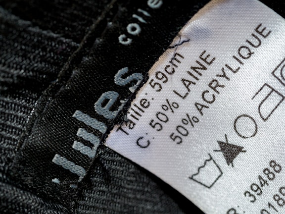 A close-up view of a clothing label showing washing instructions and material composition. The label states the size as 59 cm and the material as 50% wool (laine) and 50% acrylic. Laundry symbols are visible, including one indicating a prohibition against bleaching.