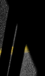 Shadows cast across a textured surface appear alongside yellow painted lines that resemble road markings. The contrast between light and dark creates an abstract, striking visual.
