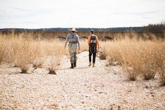 Two people wearing fishing gear walk along a dry, rocky riverbed. They carry fishing rods while surrounded by tall, dry grass. In the background, an old stone bridge stretches across the landscape, and a forested area is visible in the distance.