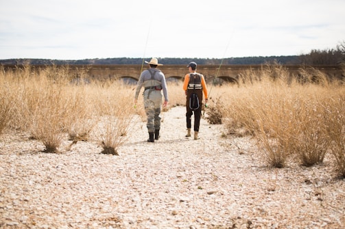 Two people wearing fishing gear walk along a dry, rocky riverbed. They carry fishing rods while surrounded by tall, dry grass. In the background, an old stone bridge stretches across the landscape, and a forested area is visible in the distance.