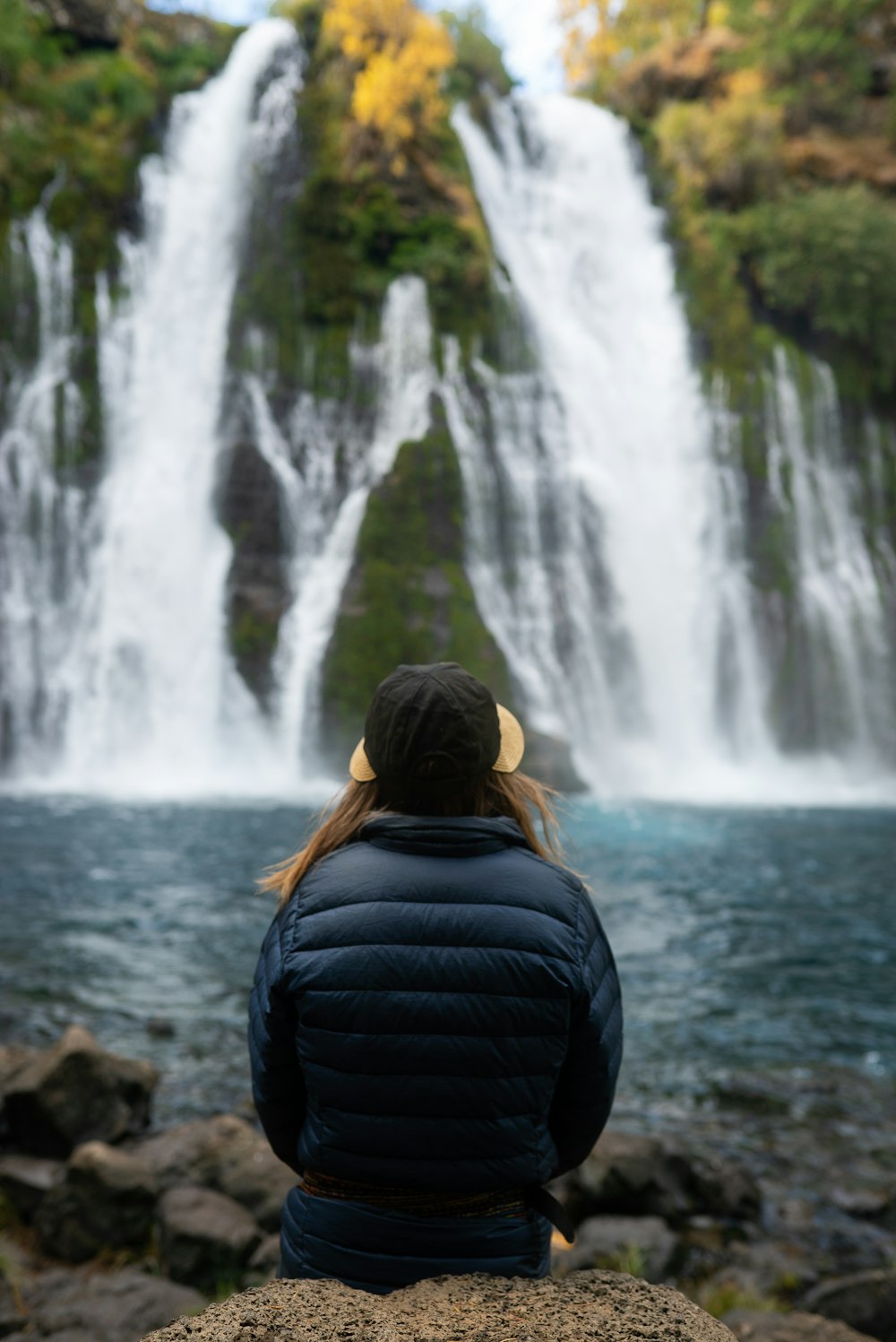 昼間、滝の近くの岩の上に座っている黒いジャケットの女性