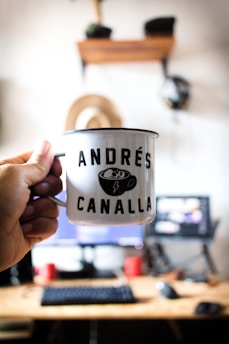 A hand holds a white metal mug with the words 'ANDRÉS CANALLA' printed on it, featuring a small graphic of a cup with a lightning bolt. The background shows a blurred home office setup with a computer monitor, keyboard, and desk. A wooden shelf with various items, including a helmet, hangs on the wall.
