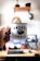 A hand holds a white metal mug with the words 'ANDRÉS CANALLA' printed on it, featuring a small graphic of a cup with a lightning bolt. The background shows a blurred home office setup with a computer monitor, keyboard, and desk. A wooden shelf with various items, including a helmet, hangs on the wall.