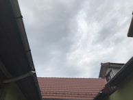 A view of rooftops with reddish-brown tiles, framed by wooden and metal guttering, and an overcast sky with grey clouds overhead.