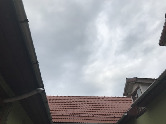 A view of rooftops with reddish-brown tiles, framed by wooden and metal guttering, and an overcast sky with grey clouds overhead.
