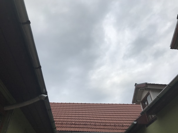 A view of rooftops with reddish-brown tiles, framed by wooden and metal guttering, and an overcast sky with grey clouds overhead.