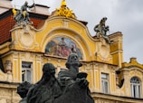 A historic architectural facade features intricate sculptures and ornate decor. The facade includes a detailed mural with figures, surrounded by embellishments in yellow and beige tones. At the forefront stands a dark-colored statue of multiple figures, adding contrast against the brighter background.