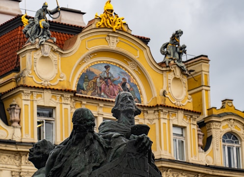 A historic architectural facade features intricate sculptures and ornate decor. The facade includes a detailed mural with figures, surrounded by embellishments in yellow and beige tones. At the forefront stands a dark-colored statue of multiple figures, adding contrast against the brighter background.
