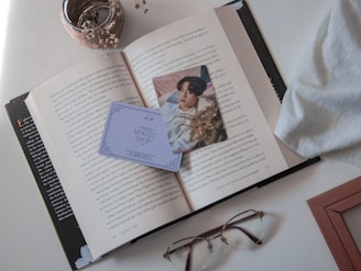 An open book with a photo card and a 'Magic Shop' card on top. A glass jar with dried flowers is placed beside the book. Nearby are a pair of glasses and a piece of white fabric.