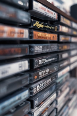 A close-up view of a collection of vintage music cassette tapes stacked together, featuring a variety of artists and music genres. The spines of the cassette cases display the names of musicians and albums in bold, colorful fonts.