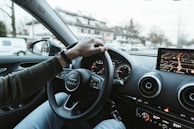 A person is driving a car with a visible navigation system on the dashboard. The hand is on the steering wheel, and houses can be seen outside through the windshield.