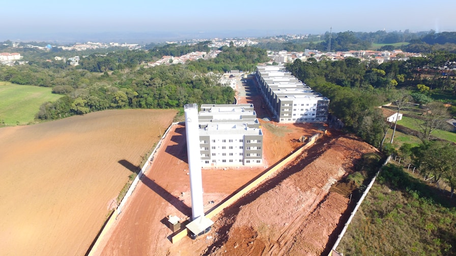 Aerial view of a construction site with several newly built buildings arranged in rows, surrounded by large areas of cleared land and forest. The site is bordered by a cityscape in the distance.