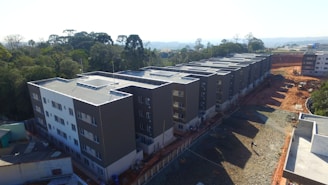 A row of newly constructed residential apartment buildings with multiple floors, aligned next to each other. The buildings have a modern design with dark gray and white colors. The surrounding area includes a construction site with red earth and gravel. Dense green trees are visible in the background, suggesting a suburban or semi-rural setting.