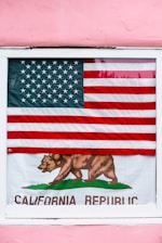 An American flag is displayed outside a window, partially covering a California Republic flag. The American flag has red and white stripes with a blue field of white stars. The California Republic flag features a bear walking on green grass and the text 'California Republic'. The background is a pink wall.