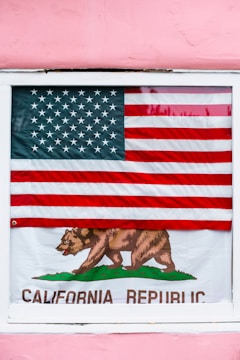 An American flag is displayed outside a window, partially covering a California Republic flag. The American flag has red and white stripes with a blue field of white stars. The California Republic flag features a bear walking on green grass and the text 'California Republic'. The background is a pink wall.