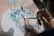 A person is holding a small paintbrush and applying vibrant colors onto a mixing palette. Various shades of blue, pink, red, and green are spread across the palette, suggesting preparation for a creative painting session.