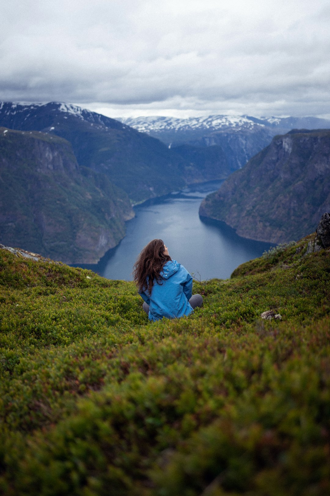 woman in blue jacket sitting on green grass field near lake during daytime