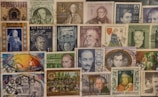 A collection of various vintage postage stamps, mostly from Austria. The stamps feature portraits of notable figures, historic landmarks, and artistic illustrations. Some stamps have been postmarked, indicating they have been used. The arrangement is collage-like, creating a busy and colorful display.