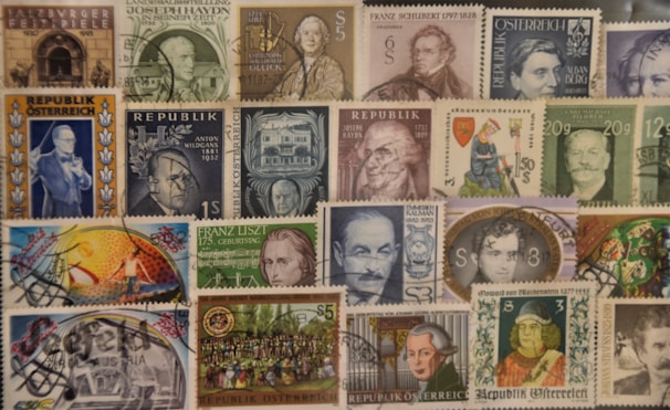 A collection of various vintage postage stamps, mostly from Austria. The stamps feature portraits of notable figures, historic landmarks, and artistic illustrations. Some stamps have been postmarked, indicating they have been used. The arrangement is collage-like, creating a busy and colorful display.