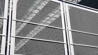 A close-up view of a metal grid or fence, with geometric patterns formed by intersecting lines. The structure appears sturdy, with a metallic sheen that reflects light. Behind the grid, industrial elements like beams or trusses are faintly visible, adding depth to the composition.
