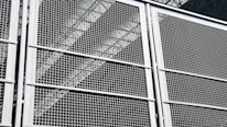 A close-up view of a metal grid or fence, with geometric patterns formed by intersecting lines. The structure appears sturdy, with a metallic sheen that reflects light. Behind the grid, industrial elements like beams or trusses are faintly visible, adding depth to the composition.