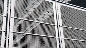 A close-up view of a metal grid or fence, with geometric patterns formed by intersecting lines. The structure appears sturdy, with a metallic sheen that reflects light. Behind the grid, industrial elements like beams or trusses are faintly visible, adding depth to the composition.