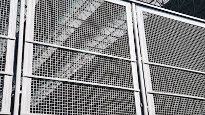 A close-up view of a metal grid or fence, with geometric patterns formed by intersecting lines. The structure appears sturdy, with a metallic sheen that reflects light. Behind the grid, industrial elements like beams or trusses are faintly visible, adding depth to the composition.