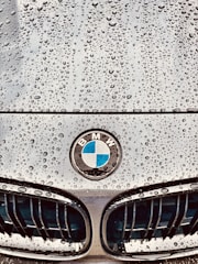 Raindrops are scattered across the smooth surface of a car hood, with a BMW emblem prominently displayed in the center. The grille design is visible below the emblem, showing sleek lines and a polished finish.