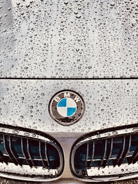 Raindrops are scattered across the smooth surface of a car hood, with a BMW emblem prominently displayed in the center. The grille design is visible below the emblem, showing sleek lines and a polished finish.