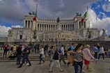 A large neoclassical monument with grand columns and sculptures, featuring two Italian flags. Numerous people are walking and gathering in front of the monument, some taking photos. The sky is partly cloudy, adding a dynamic backdrop to the scene.