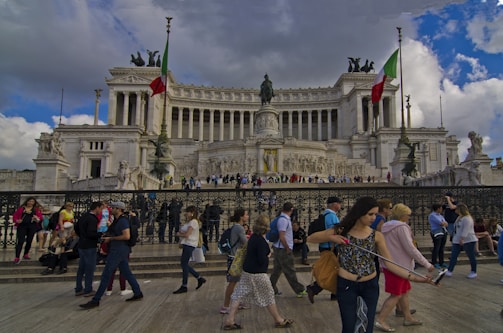 A large neoclassical monument with grand columns and sculptures, featuring two Italian flags. Numerous people are walking and gathering in front of the monument, some taking photos. The sky is partly cloudy, adding a dynamic backdrop to the scene.