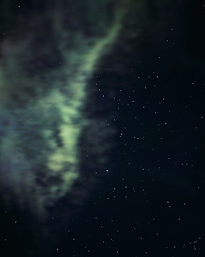 Swirling greenish aurora set against a backdrop of a starry night sky. Wispy clouds mix with scattered stars, creating a celestial scene.