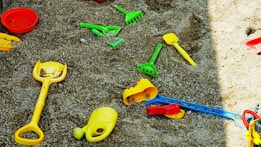 Various colorful plastic sand toys, including shovels and rakes, are scattered across a sandy surface. The toys are mostly in primary colors such as yellow, green, and red.