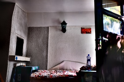 A cozy interior space with textured walls featuring a small bed covered with a colorful patterned blanket. A black wall-mounted lamp and a small framed picture adorn the walls. A clear plastic water bottle sits on a surface next to the bed, and light filters in through a nearby window, casting shadows on the walls.
