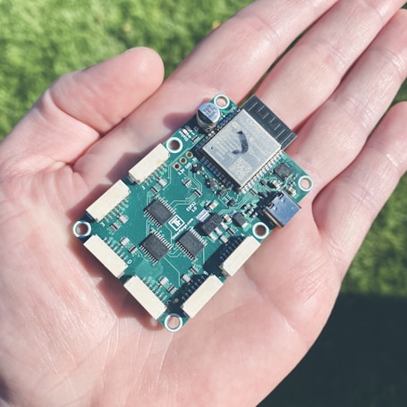 A small printed circuit board with various electronic components is resting on the palm of a hand. The circuit board features several chips, connectors, and capacitors, with intricate soldering and wiring visible.