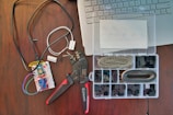 A workspace featuring electronic components including a breadboard with wires connected, a wire cutter or crimping tool, and an organized plastic box with various electronic connectors. Part of a laptop keyboard can also be seen in the corner.