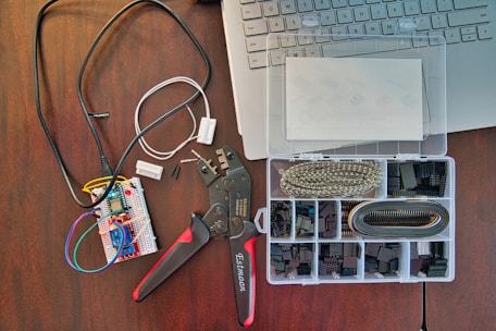 A workspace featuring electronic components including a breadboard with wires connected, a wire cutter or crimping tool, and an organized plastic box with various electronic connectors. Part of a laptop keyboard can also be seen in the corner.
