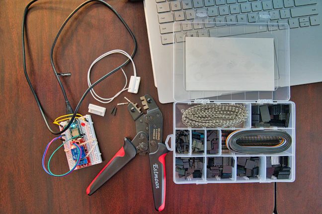 A workspace featuring electronic components including a breadboard with wires connected, a wire cutter or crimping tool, and an organized plastic box with various electronic connectors. Part of a laptop keyboard can also be seen in the corner.