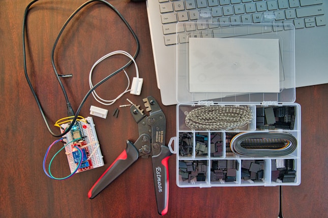 A workspace featuring electronic components including a breadboard with wires connected, a wire cutter or crimping tool, and an organized plastic box with various electronic connectors. Part of a laptop keyboard can also be seen in the corner.